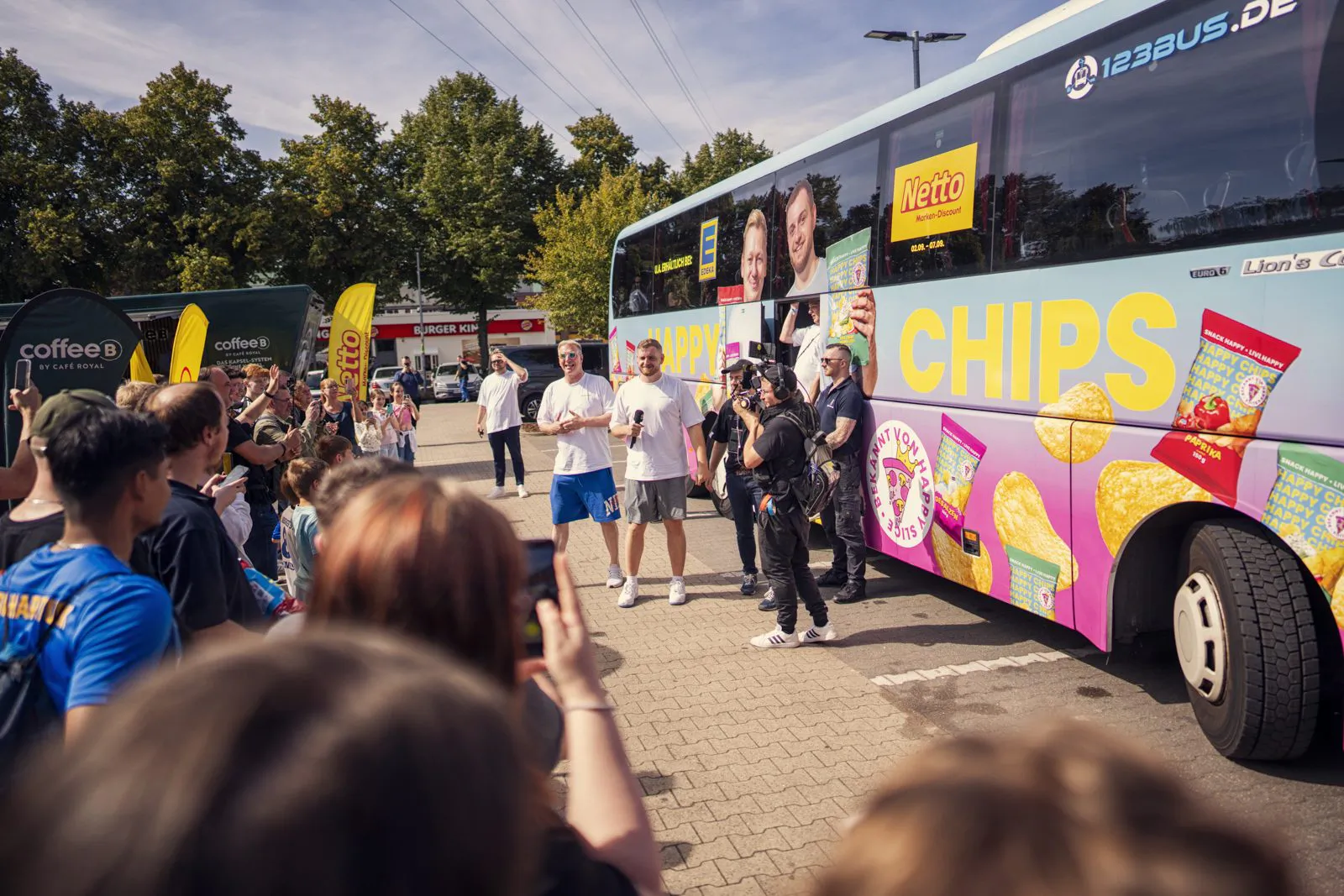 Menschen und Kamerateam vor einem Netto-Happy-Chips-Bus auf einem sonnigen Veranstaltungsplatz.