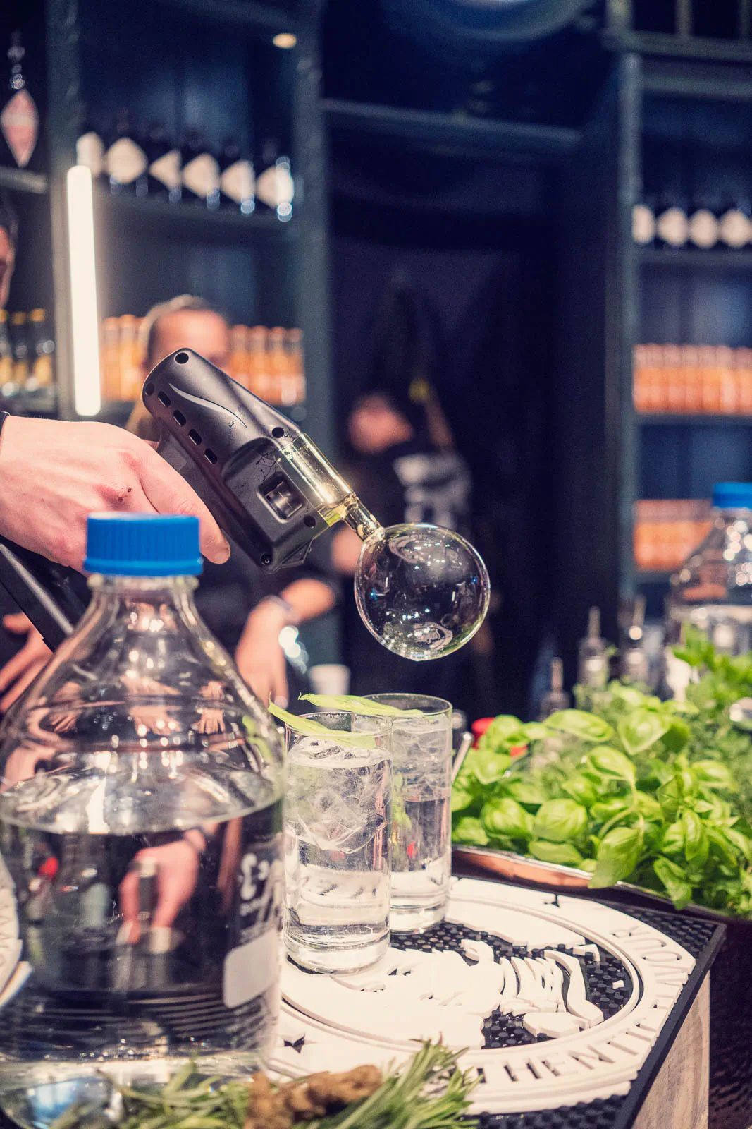An einer dunklen Bar hält eine Person ein Gerät mit Glaskugel über Drinks mit Eis und Limette, daneben frische Kräuter.