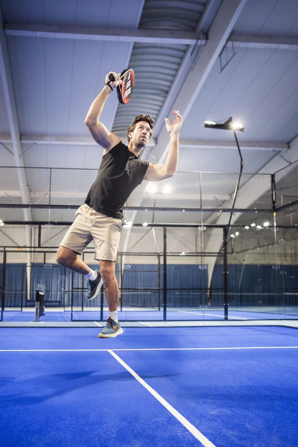 Mann mit Padelschläger springt zum Überkopfschlag auf einem blauen Padelplatz in einer hell beleuchteten Halle.