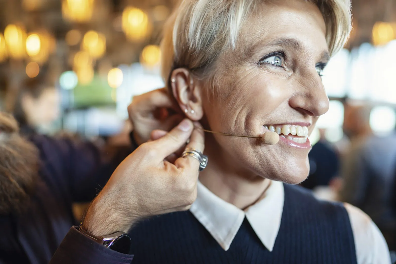 Einer lächelnden Frau wird bei warmem Licht in einem Innenraum ein Headset-Mikrofon am Ohr befestigt.