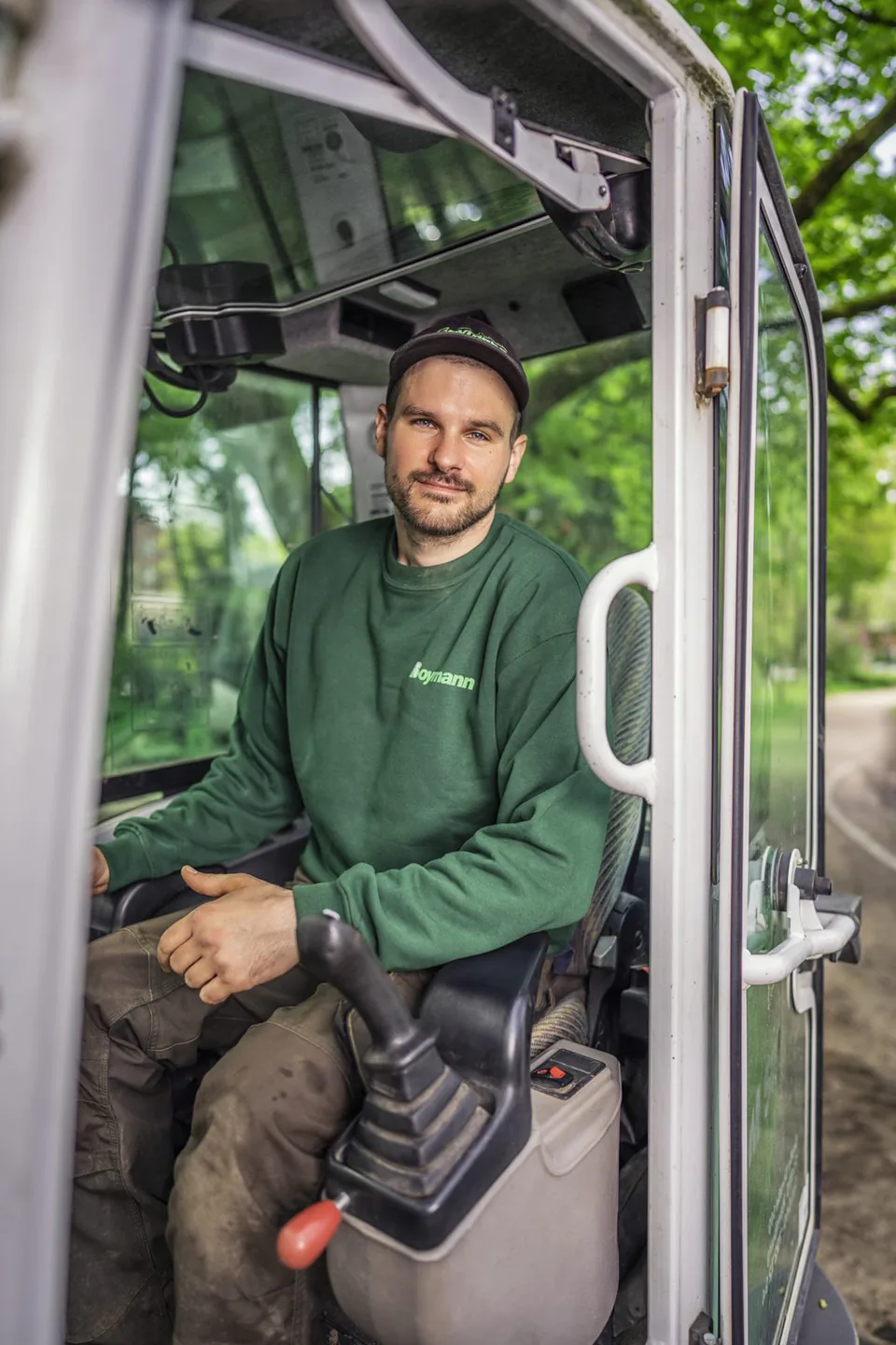 Mann im grünen Boymann-Pullover sitzt bei Tageslicht in der offenen Kabine einer Baumaschine, umgeben von Bäumen.