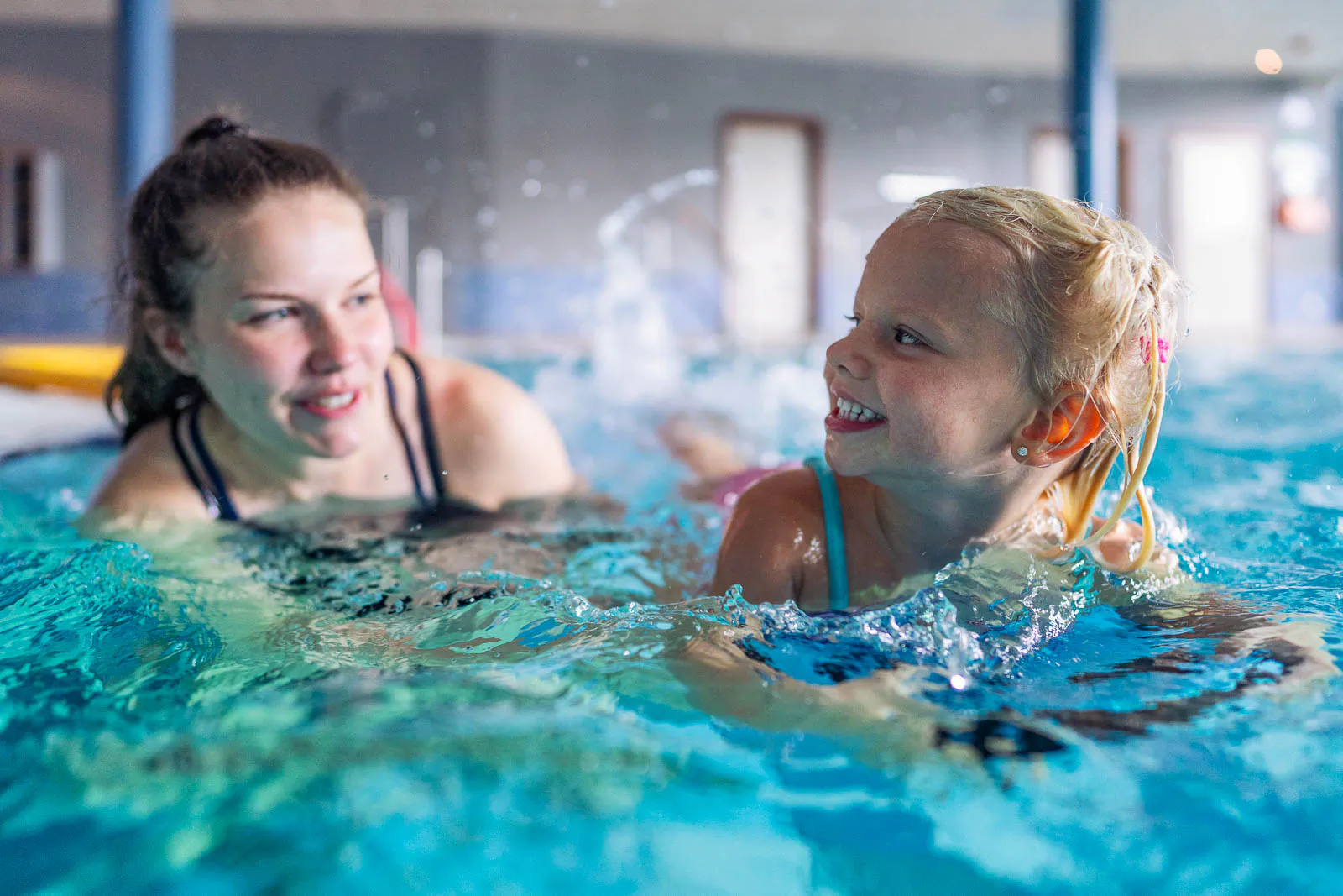 Eine Frau begleitet ein lächelndes Mädchen beim Schwimmen im türkisblauen Wasser eines hellen Hallenbads.