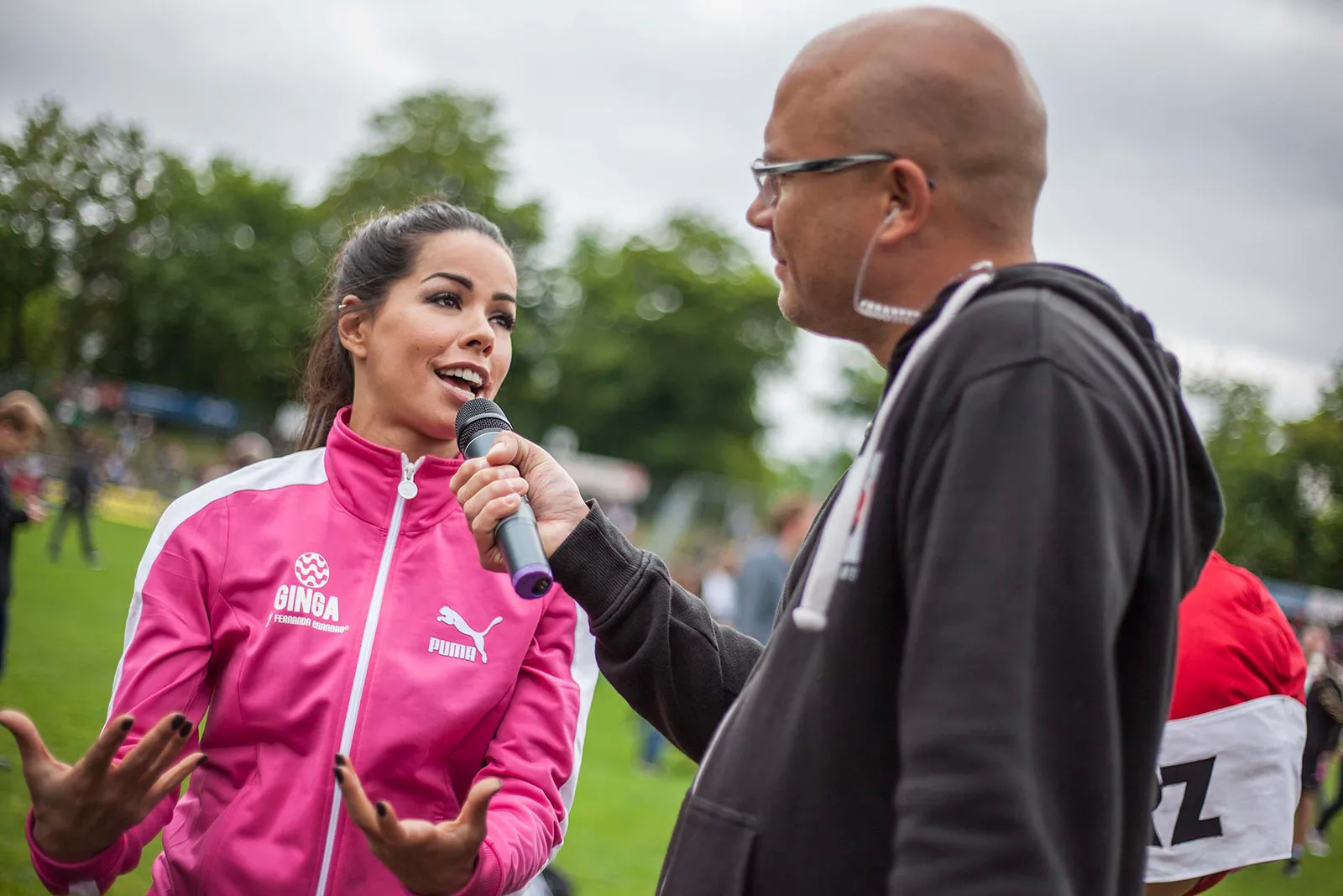 Frau in pinker Sportjacke spricht auf einem Rasenplatz in ein Mikrofon, das ein Mann mit Brille hält.
