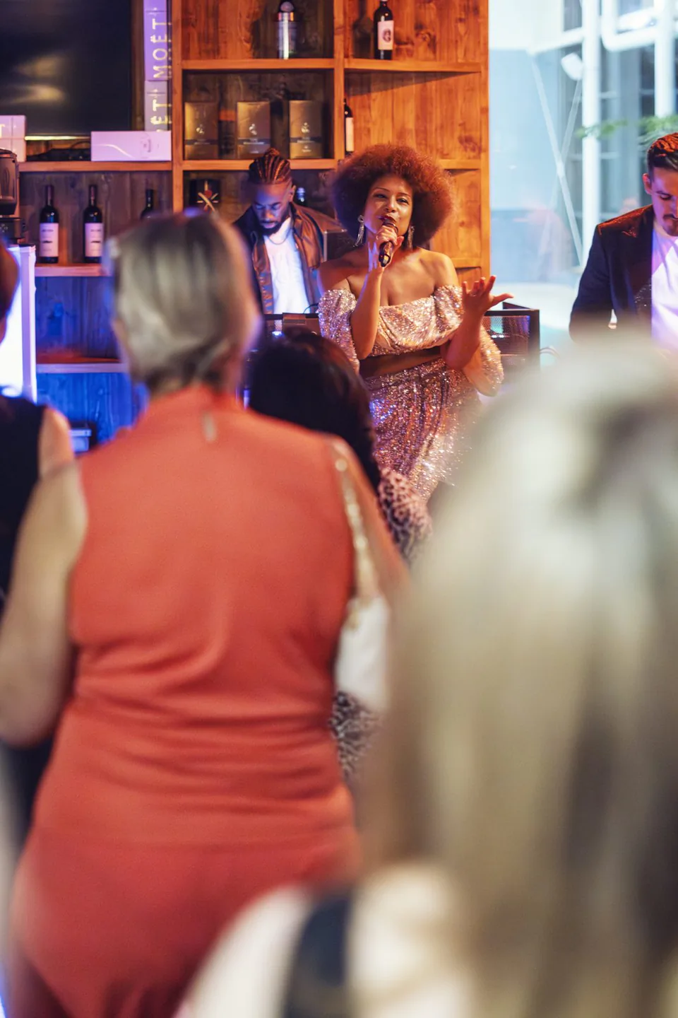 Sängerin im glitzernden Kleid singt mit Mikrofon vor Gästen, begleitet von Musikern vor hölzernen Weinregalen.