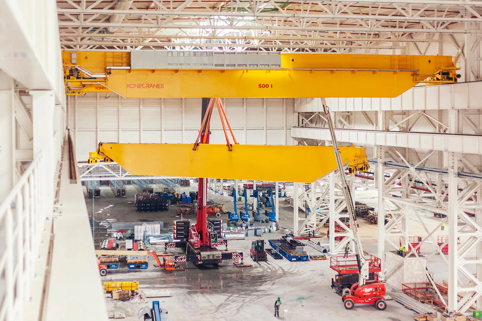 Gelber KONECRANES-Brückenkran hebt einen Kranträger an roten Gurten in einer hellen Industriehalle.