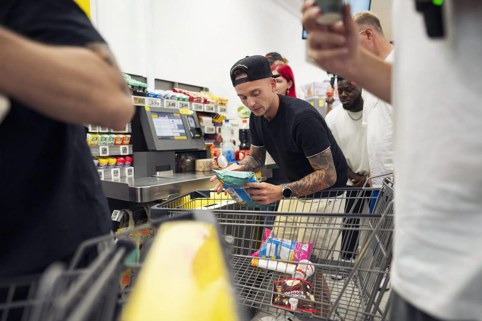 Ein tätowierter Mann mit schwarzer Kappe hält Chipstüten an einer hell beleuchteten Supermarktkasse neben einem Einkaufswagen.