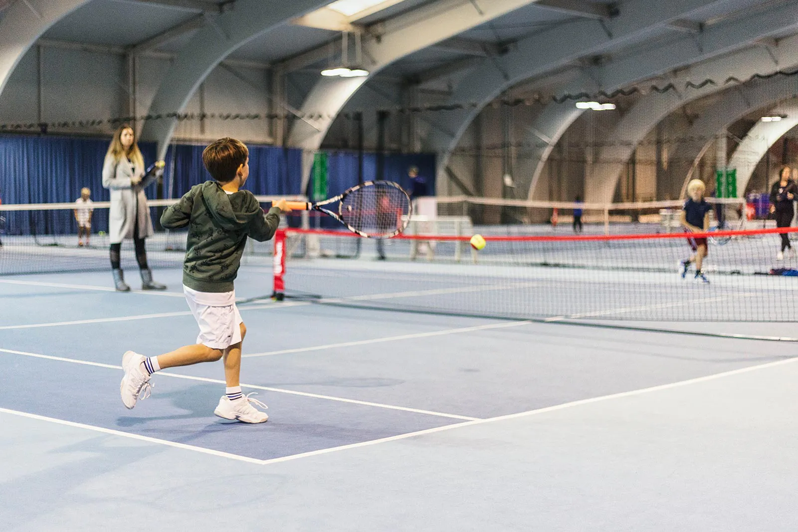 Kind schlägt einen Tennisball mit dem Schläger über ein niedriges Netz in einer hellen Tennishalle beim Wilson Kids Cup.