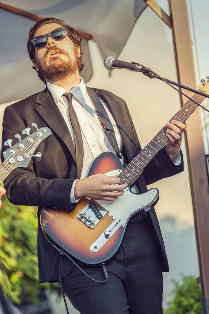Bärtiger Musiker mit Sonnenbrille und Anzug spielt E-Gitarre vor einem Mikrofon im warmen Tageslicht.