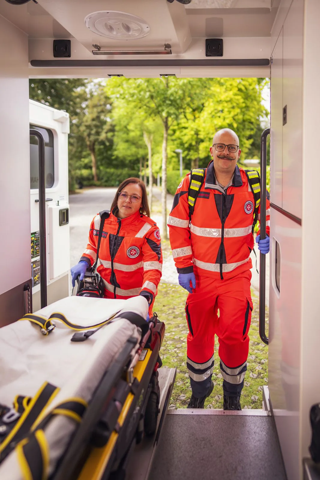 Zwei Johanniter-Rettungskräfte in roter Einsatzkleidung schieben bei Tageslicht eine Trage in einen Rettungswagen.