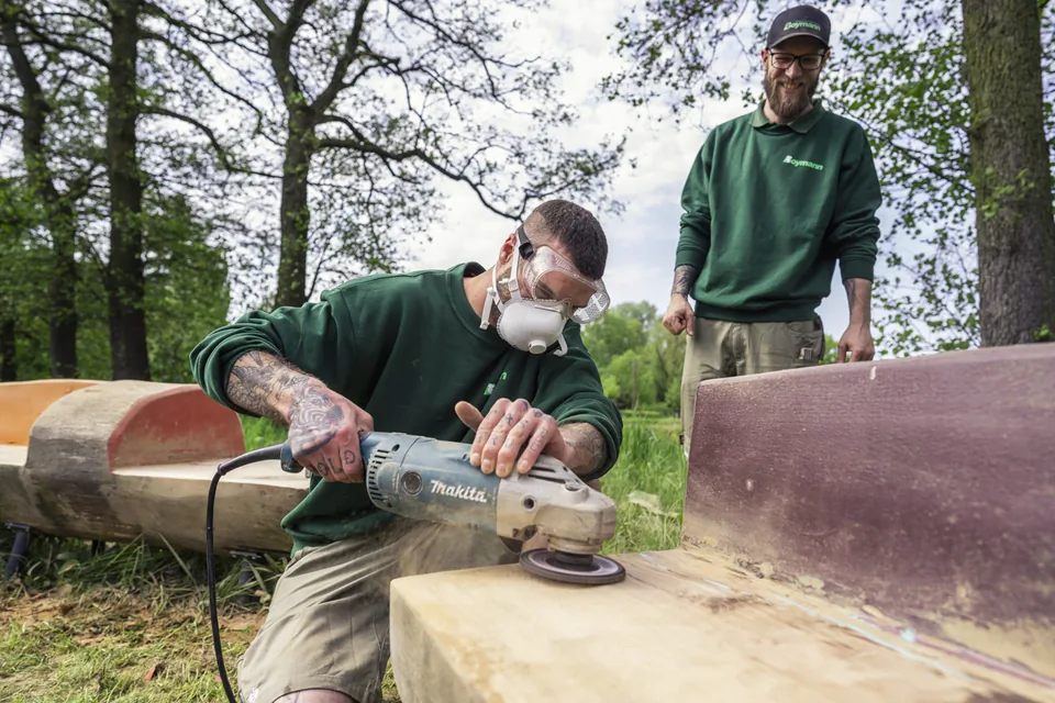 Mann mit Schutzbrille und Atemmaske schleift eine Holzbank im Freien, ein Kollege schaut zu.