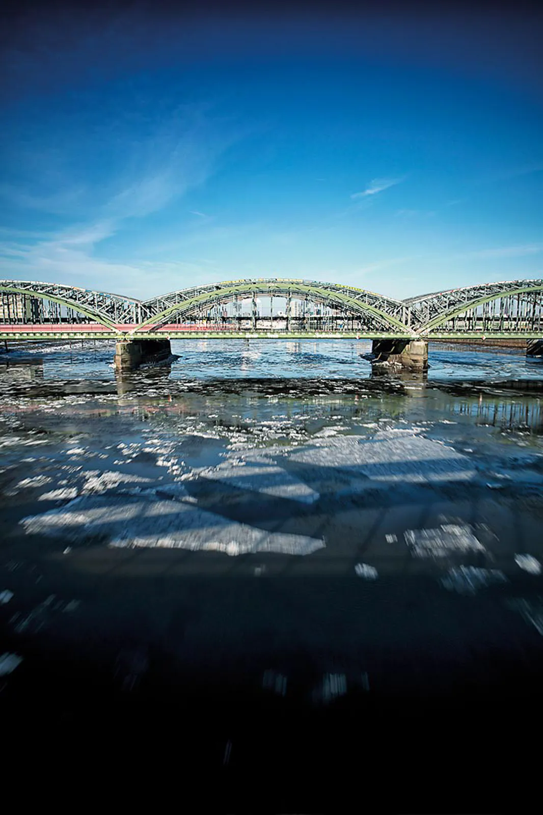 Grüne Stahlbogenbrücke über einem Fluss mit treibenden Eisschollen unter blauem Himmel im Sonnenlicht.