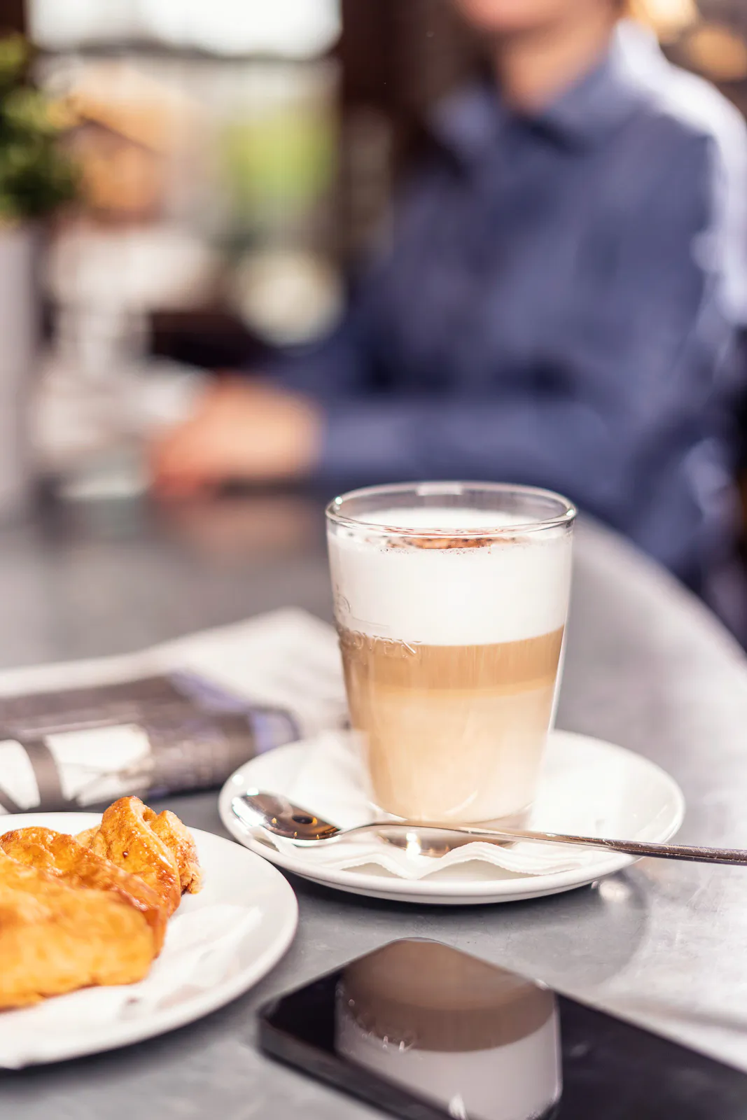 Latte macchiato im Glas, Croissant und Smartphone auf einem Cafétisch, dahinter eine unscharfe Person im Tageslicht.
