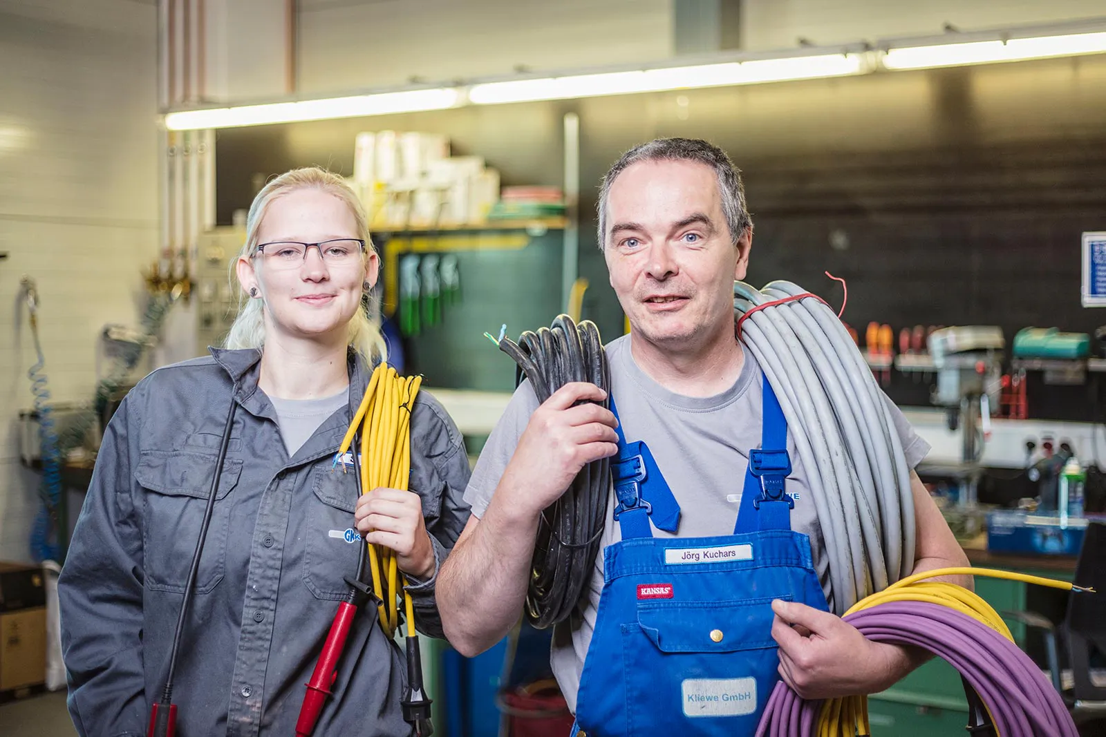Zwei Personen in Arbeitskleidung halten Kabelbündel in einer Werkstatt unter hellem Leuchtstofflicht.