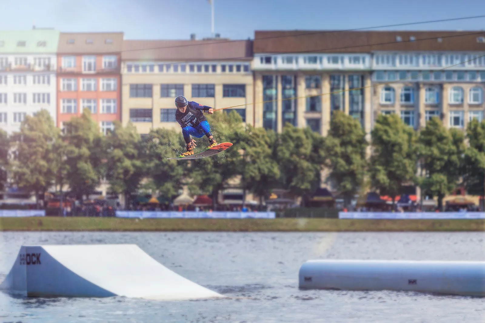 Wakeboarder springt am Seil über eine Rampe auf dem Wasser, vor sonnenbeleuchteten Gebäuden und Bäumen.
