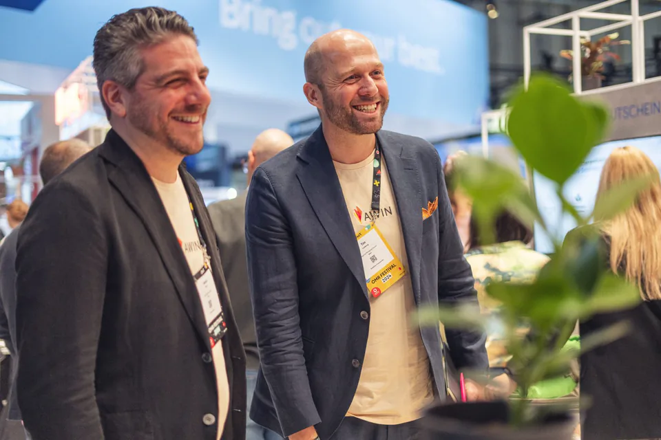 Zwei lachende Männer mit Awin-Shirts, Sakkos und Messeausweisen an einem hell beleuchteten Messestand.