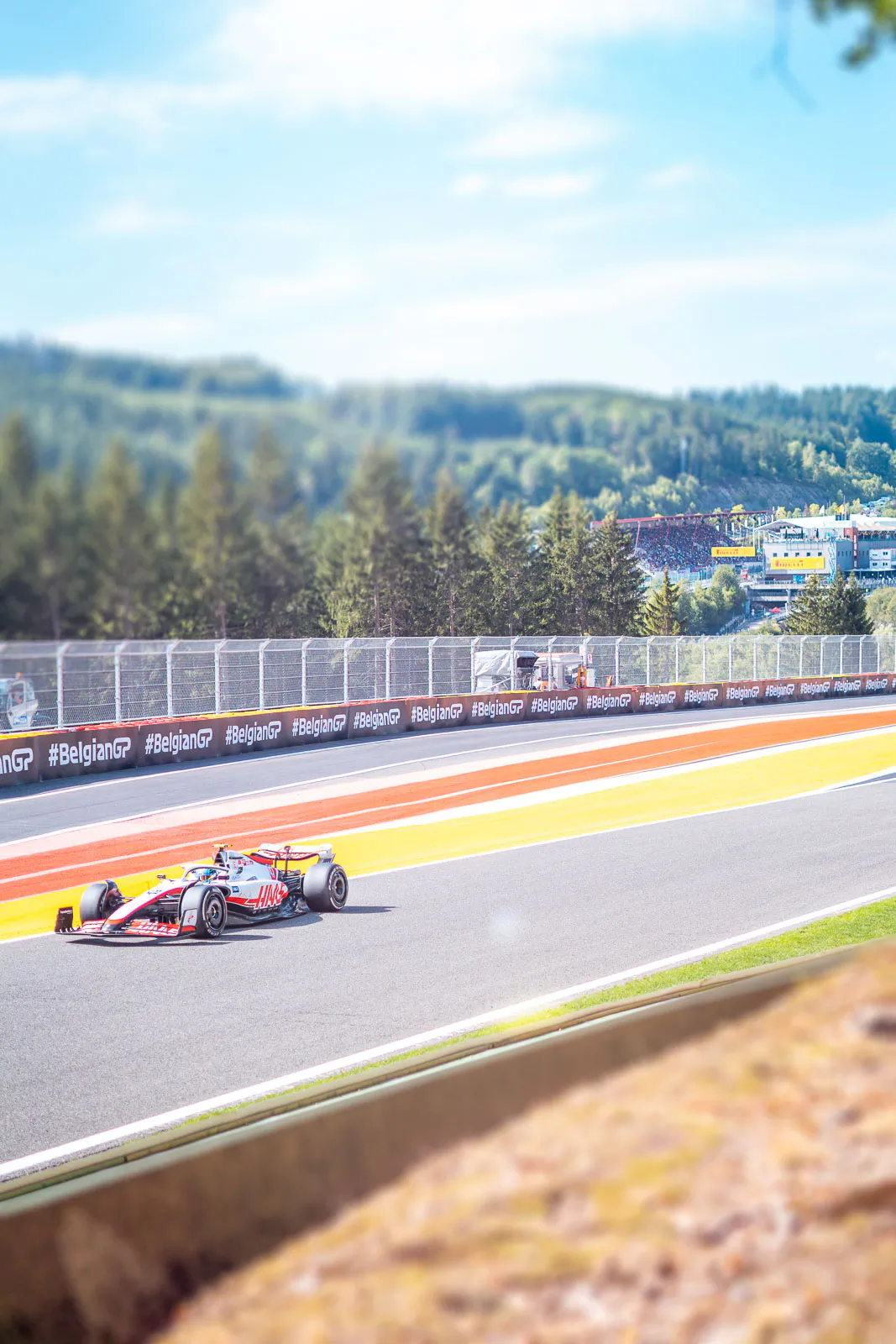 Haas-Formel-1-Wagen auf sonniger Rennstrecke mit rot-gelben Randstreifen, Fangzaun und bewaldeten Hügeln.