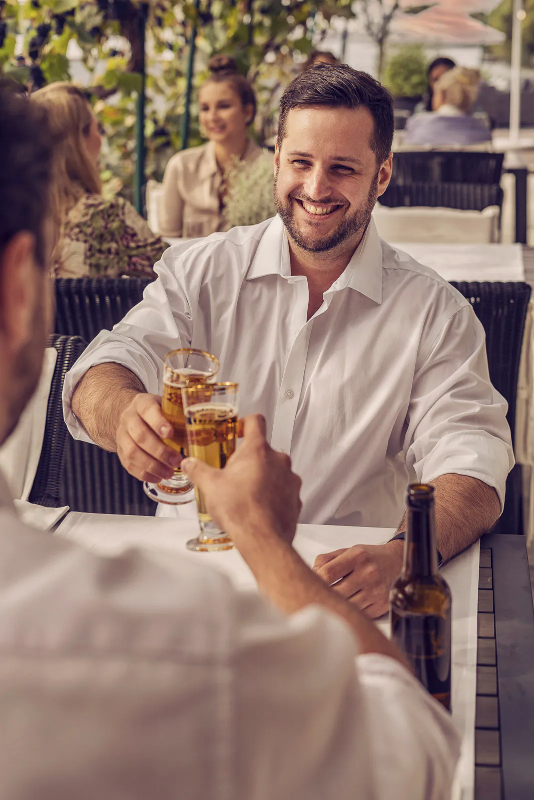 Zwei Männer in weißen Hemden stoßen auf einer sonnigen Beachclub-Terrasse mit Biergläsern an.