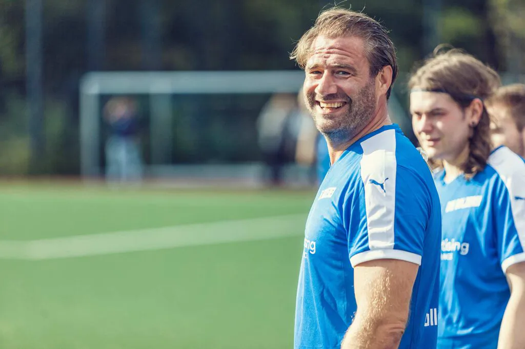 Lächelnder Mann im blau-weißen Fußballtrikot vor weiteren Spielern auf einem sonnigen Kunstrasenplatz.