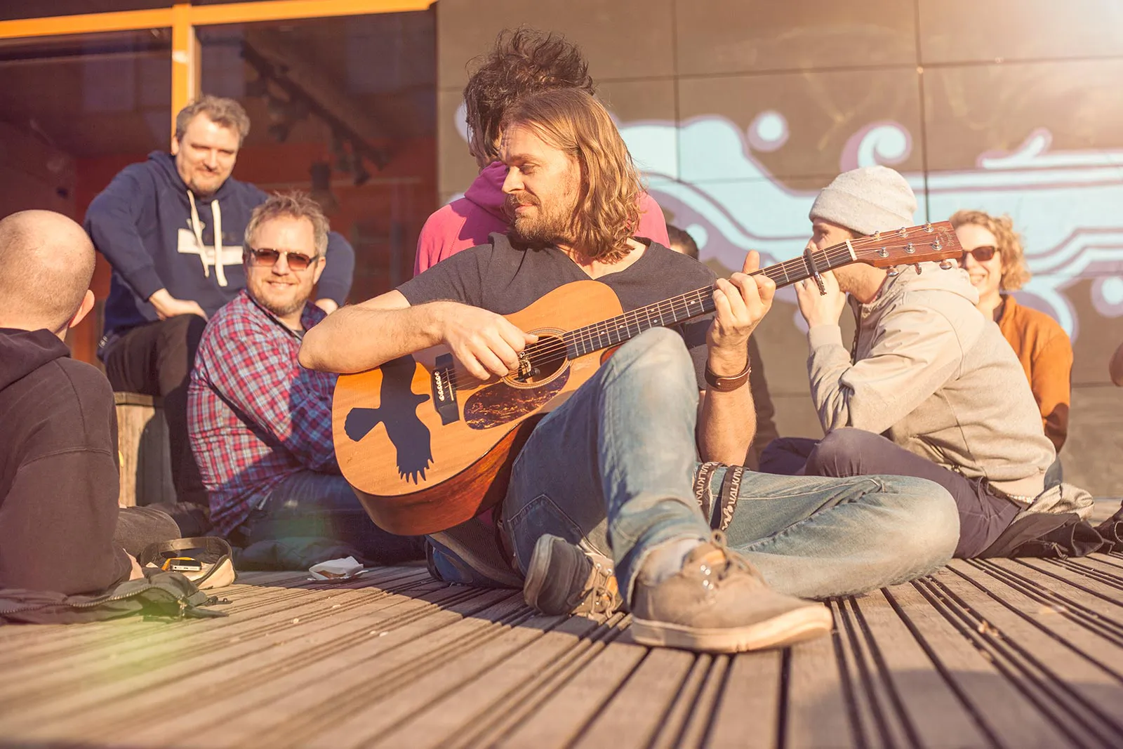 Mann spielt Akustikgitarre auf einer sonnigen Holzterrasse, umgeben von sitzenden Menschen.