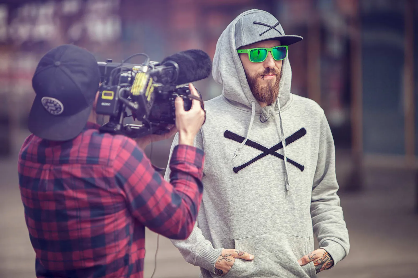 Kameramann filmt Dennis Grundt im grauen Kapuzenpullover mit grüner Sonnenbrille vor einem Gebäude.