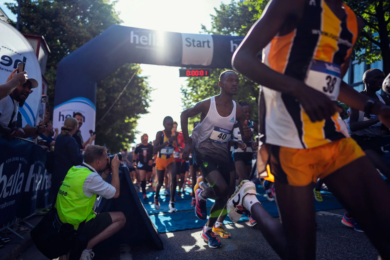 Läufer passieren im Gegenlicht den Startbogen des Hella Halbmarathons, ein Fotograf kniet am Streckenrand.