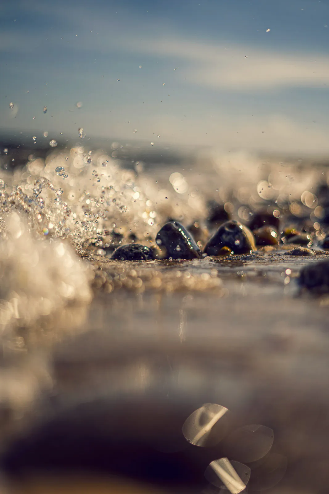 Wellen spritzen über dunkle Kiesel am Strand, Wassertropfen glitzern im warmen Sonnenlicht.