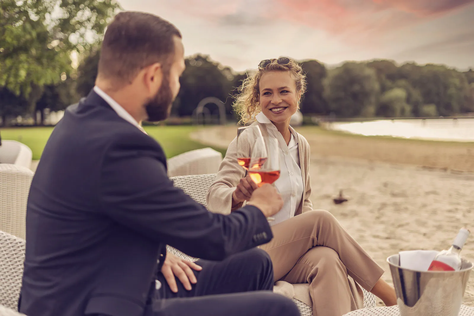 Mann und lächelnde Frau stoßen mit Weingläsern in einer Strandlounge am See im warmen Abendlicht an.