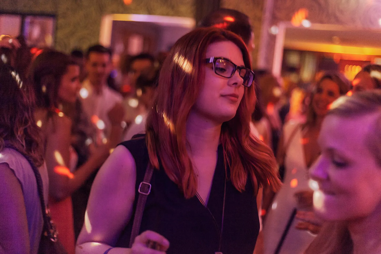 Frau mit Brille und schwarzem Oberteil zwischen Partygästen im Innenraum, beleuchtet von bunten Disco-Lichtern.