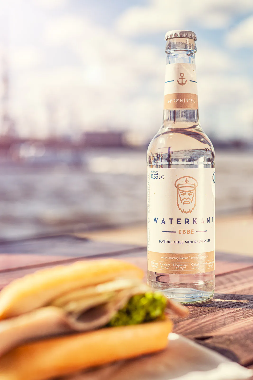 Waterkant-Mineralwasserflasche auf einem Holztisch am Hafen, Fischbrötchen im Vordergrund, warmes Sonnenlicht.