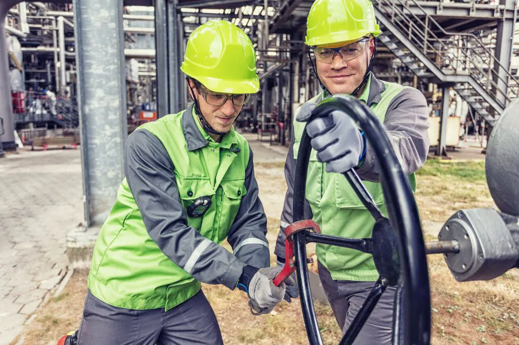Zwei Arbeiter mit grünen Helmen bedienen ein großes Handrad zwischen Rohrleitungen der Raffinerie Heide.