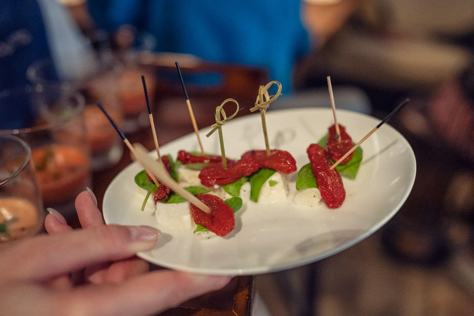 Hände halten einen weißen Teller mit aufgespießten Häppchen aus Käse, Basilikum und getrockneten Tomaten.