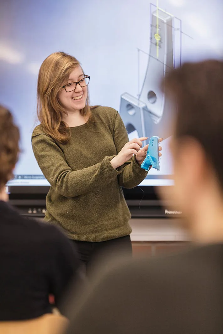 Eine Frau mit Brille erklärt im Klassenraum ein blaues Bauteil vor einer projizierten technischen Zeichnung.