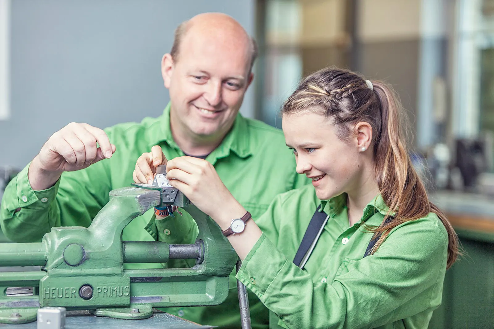 Junge Frau und Mann in grüner Arbeitskleidung bearbeiten gemeinsam ein Werkstück am Schraubstock in einer hellen Werkstatt.