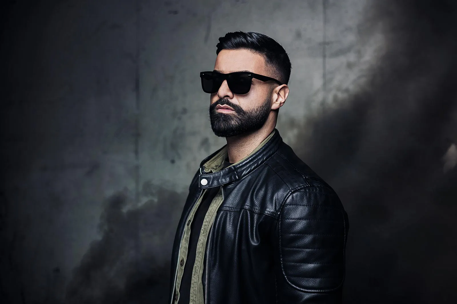Mann mit Sonnenbrille und schwarzer Lederjacke vor grauer Betonwand, seitlich beleuchtet.