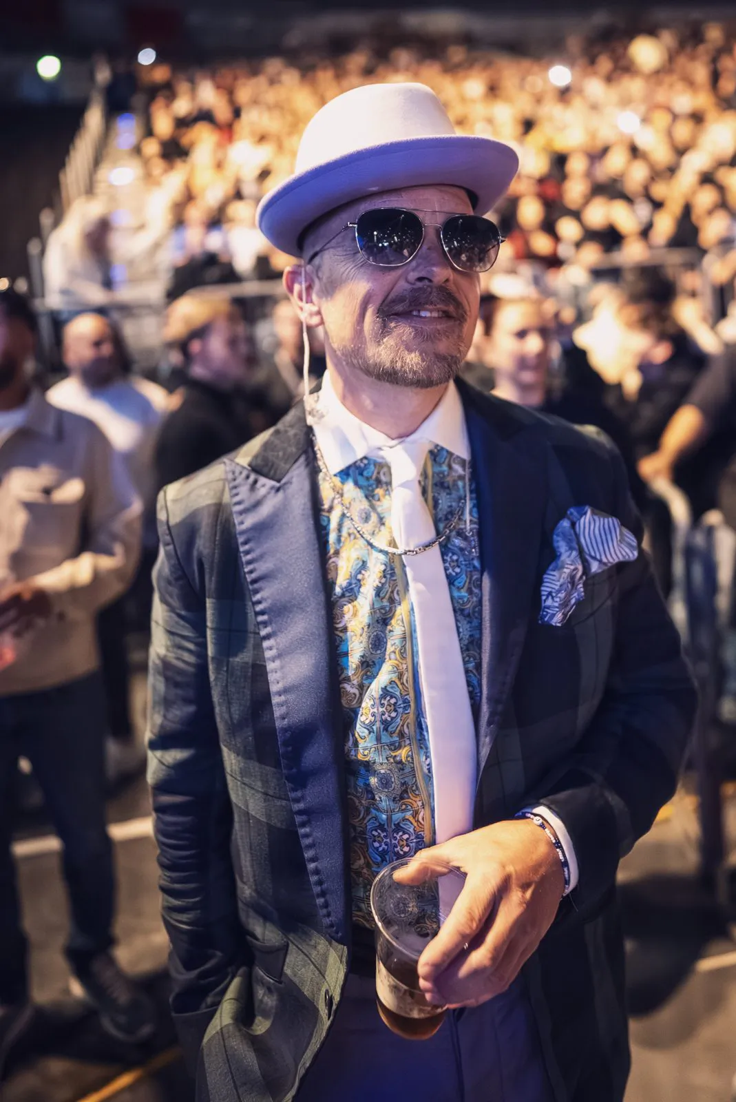 Jan Delay mit Hut, Sonnenbrille und Anzug hält einen Becher vor dichtem Konzertpublikum im warmen Licht.