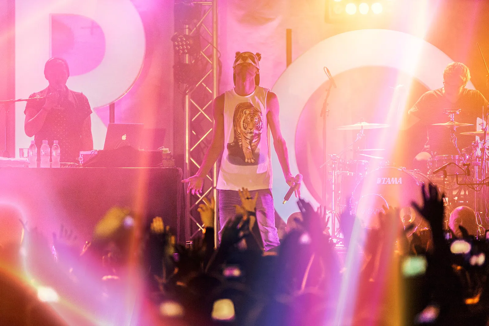 CRO mit Pandamaske und Mikrofon auf der Bühne, Band im rosa-gelben Licht und erhobene Hände im Publikum.