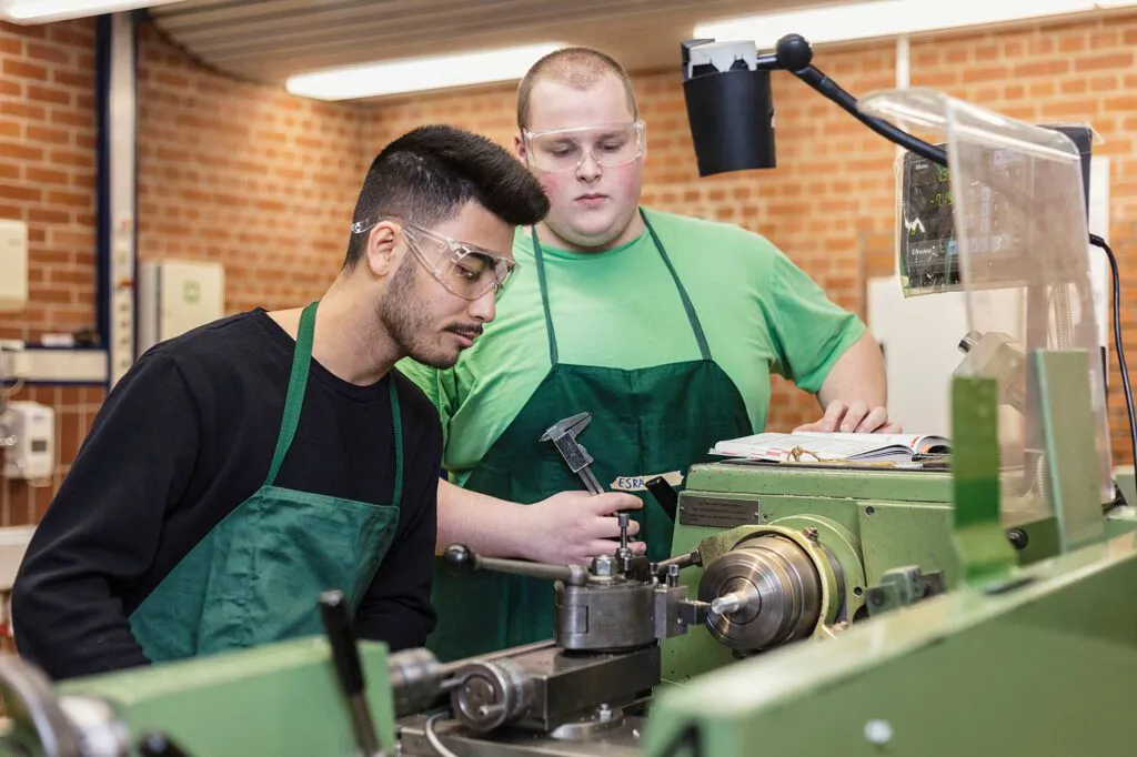 Zwei Männer in grünen Schürzen, einer mit Schutzbrille, arbeiten an einer Drehmaschine in einer hellen Werkstatt.