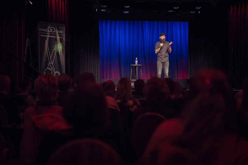 Dennis Grundt steht mit Mikrofon vor Publikum auf einer Bühne mit blau beleuchtetem Vorhang.