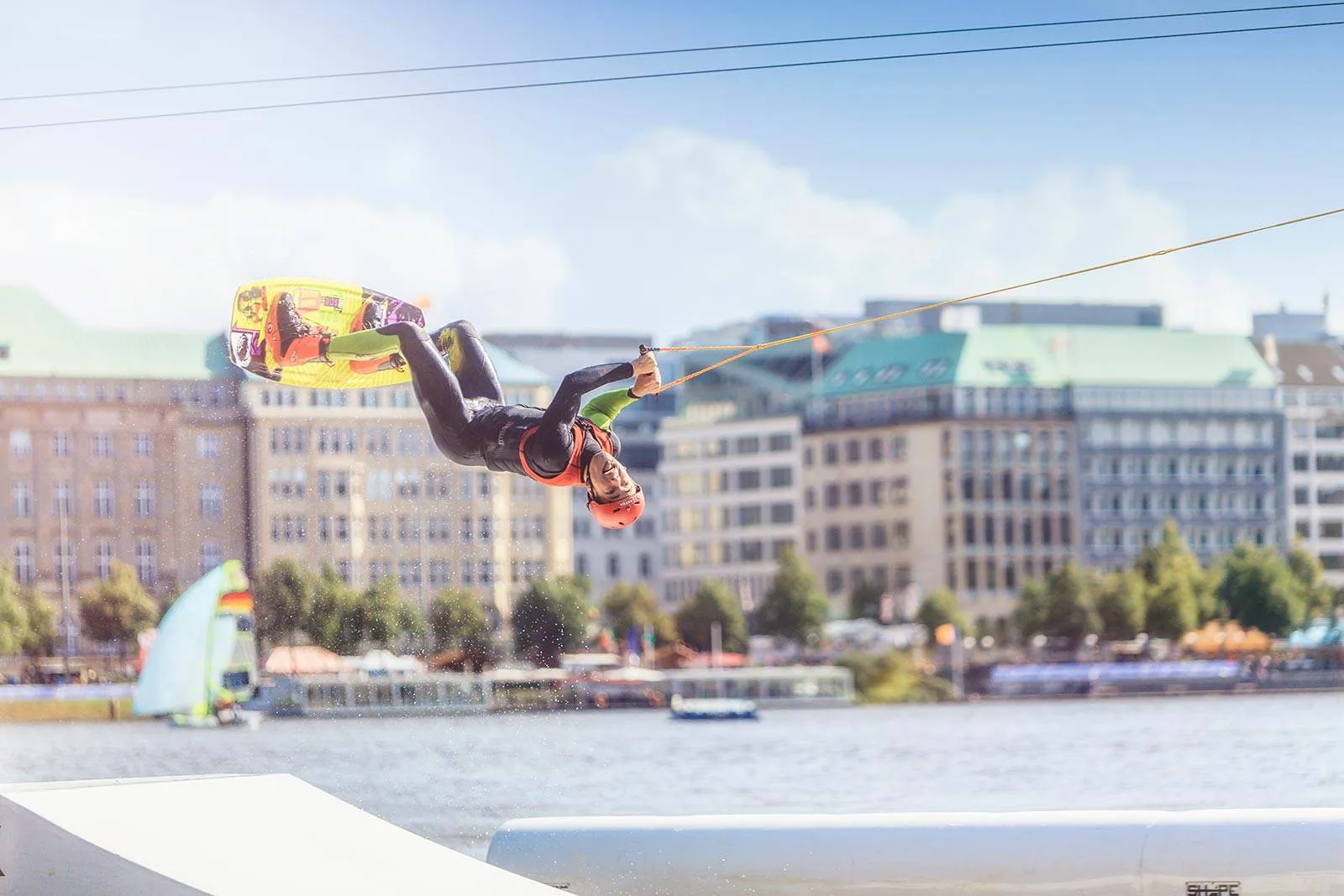 Wakeboarder kopfüber beim Sprung am Seil über sonnigem Wasser, mit buntem Board und Gebäuden am Ufer.