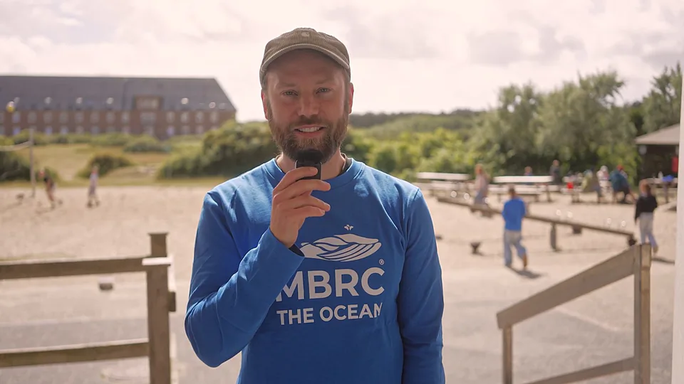 Mann mit Kappe und blauem MBRC-Shirt spricht bei Tageslicht in ein Mikrofon vor einem sandigen Außenbereich.