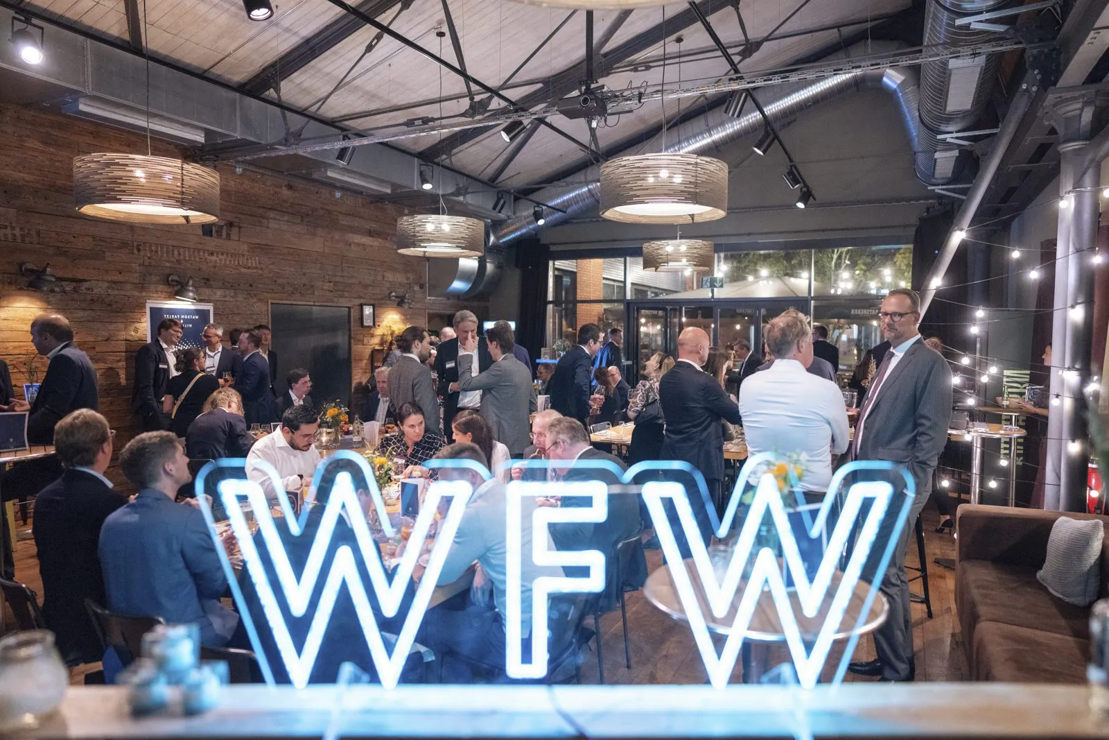 Gäste sitzen und stehen an Tischen im warm beleuchteten Eventraum, im Vordergrund leuchtet ein WFW-Schriftzug.