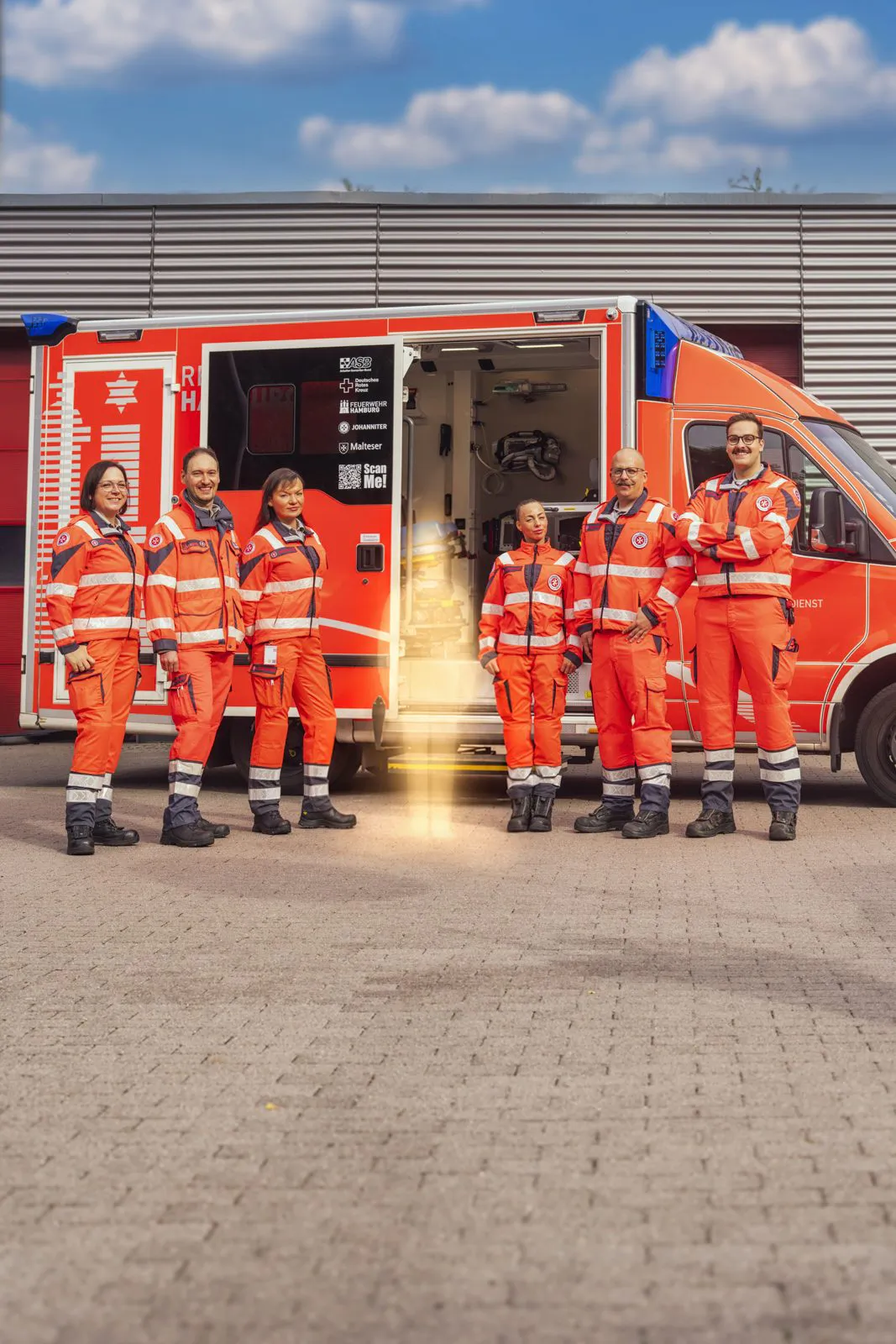 Sechs Johanniter-Rettungskräfte in orangefarbener Einsatzkleidung stehen bei Sonnenlicht vor einem offenen Rettungswagen.