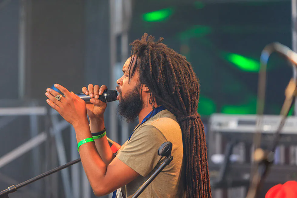 Sänger mit langen Dreadlocks singt ins Handmikrofon und gestikuliert auf einer Bühne mit grünem Licht.
