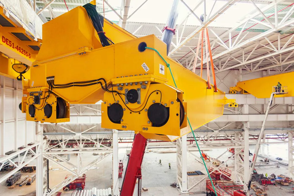 Gelbe Konecranes-Krananlage hängt an Seilen in einer hellen Industriehalle mit Stahlträgern und Hebebühnen.