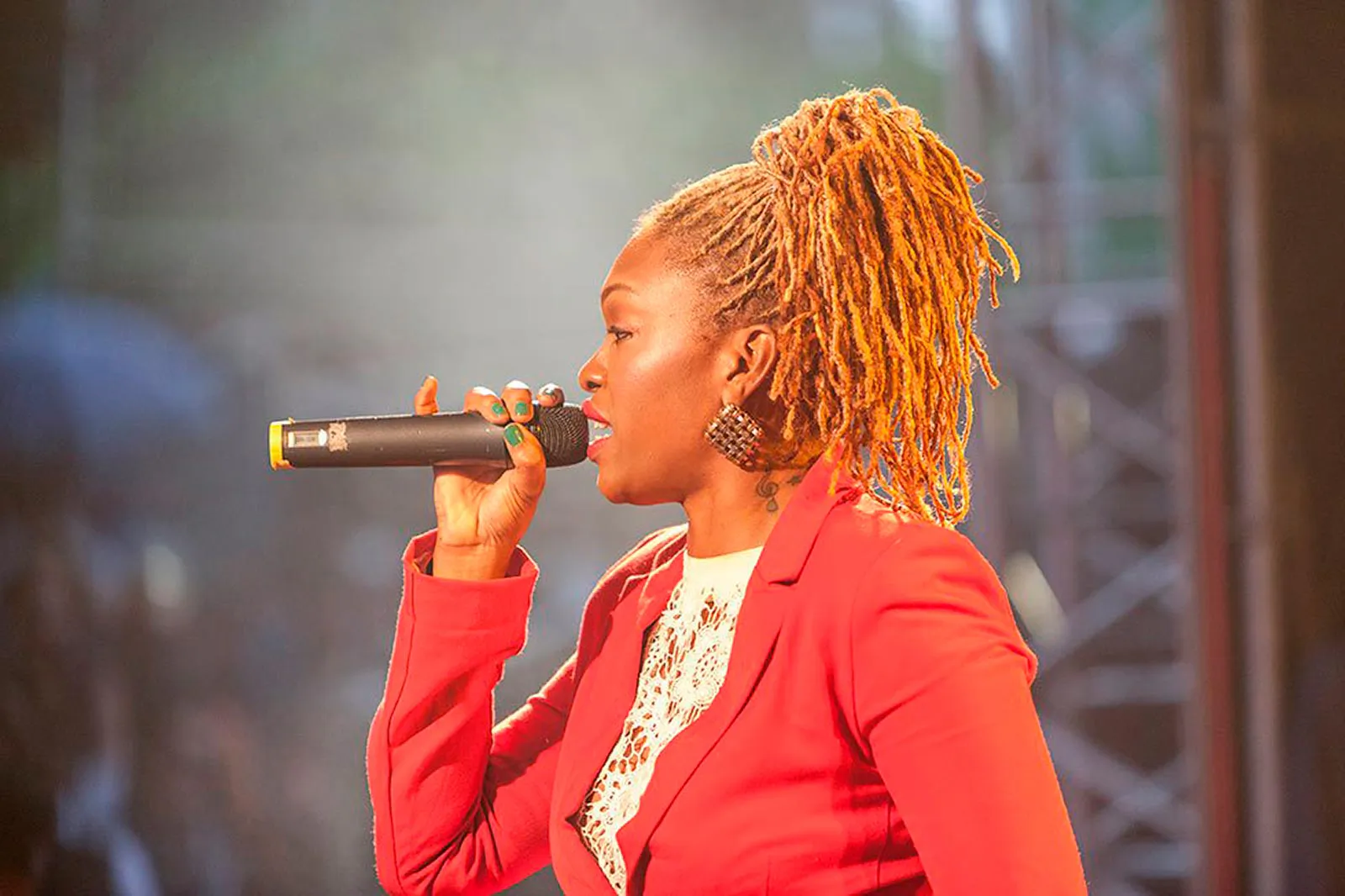 Sängerin mit hochgebundenen Dreadlocks und rotem Blazer singt im warmen Bühnenlicht beim Reggae Jam ins Mikrofon.