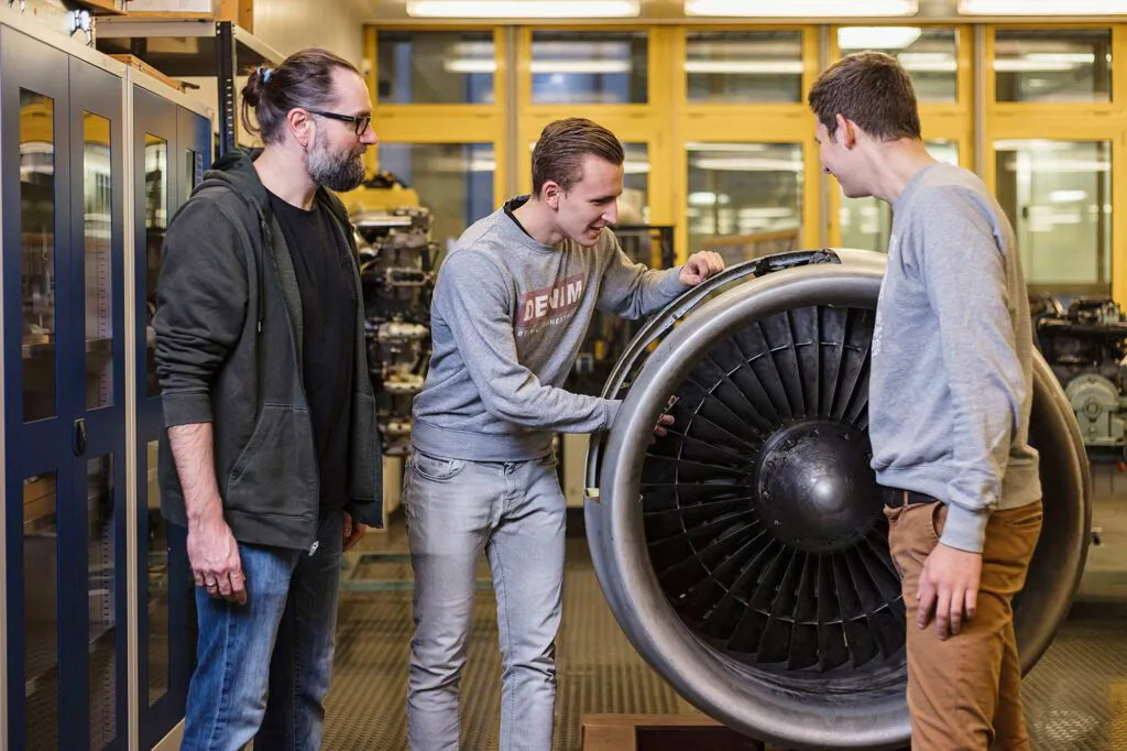 Drei Männer betrachten ein geöffnetes Flugzeugtriebwerk in einer hell beleuchteten Lehrwerkstatt.