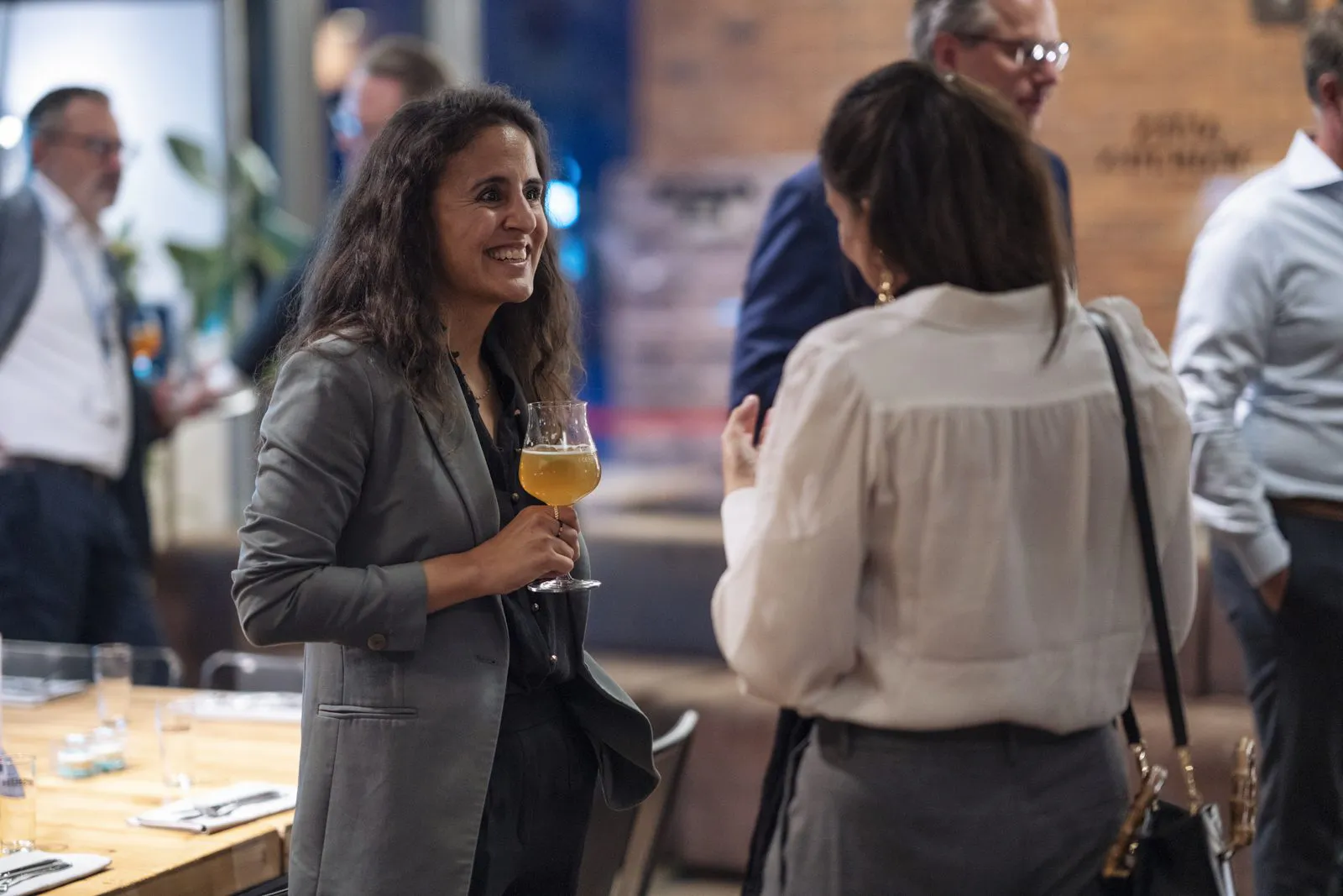 Zwei Frauen unterhalten sich beim Event in einem Innenraum; eine lächelt und hält ein Glas mit gelbem Getränk.