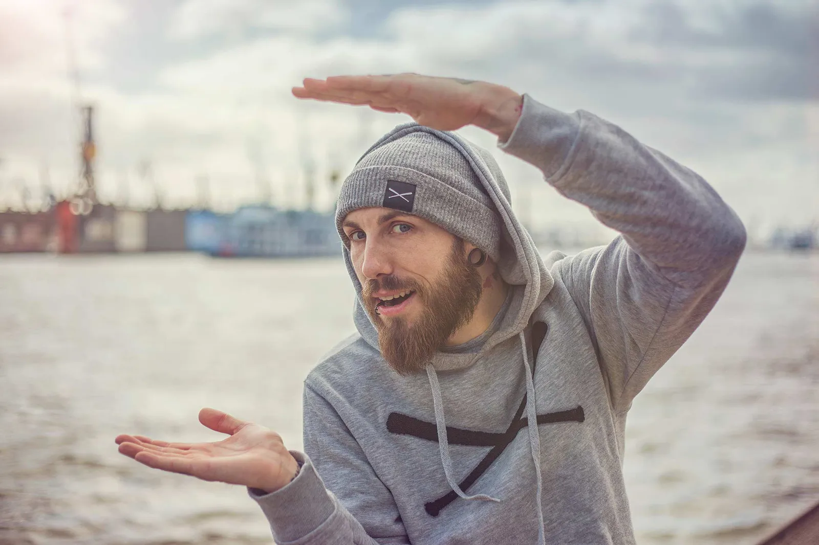 Dennis Grundt mit grauer Mütze und Hoodie gestikuliert am Hafen, eine Hand über dem Kopf, die andere nach oben offen.