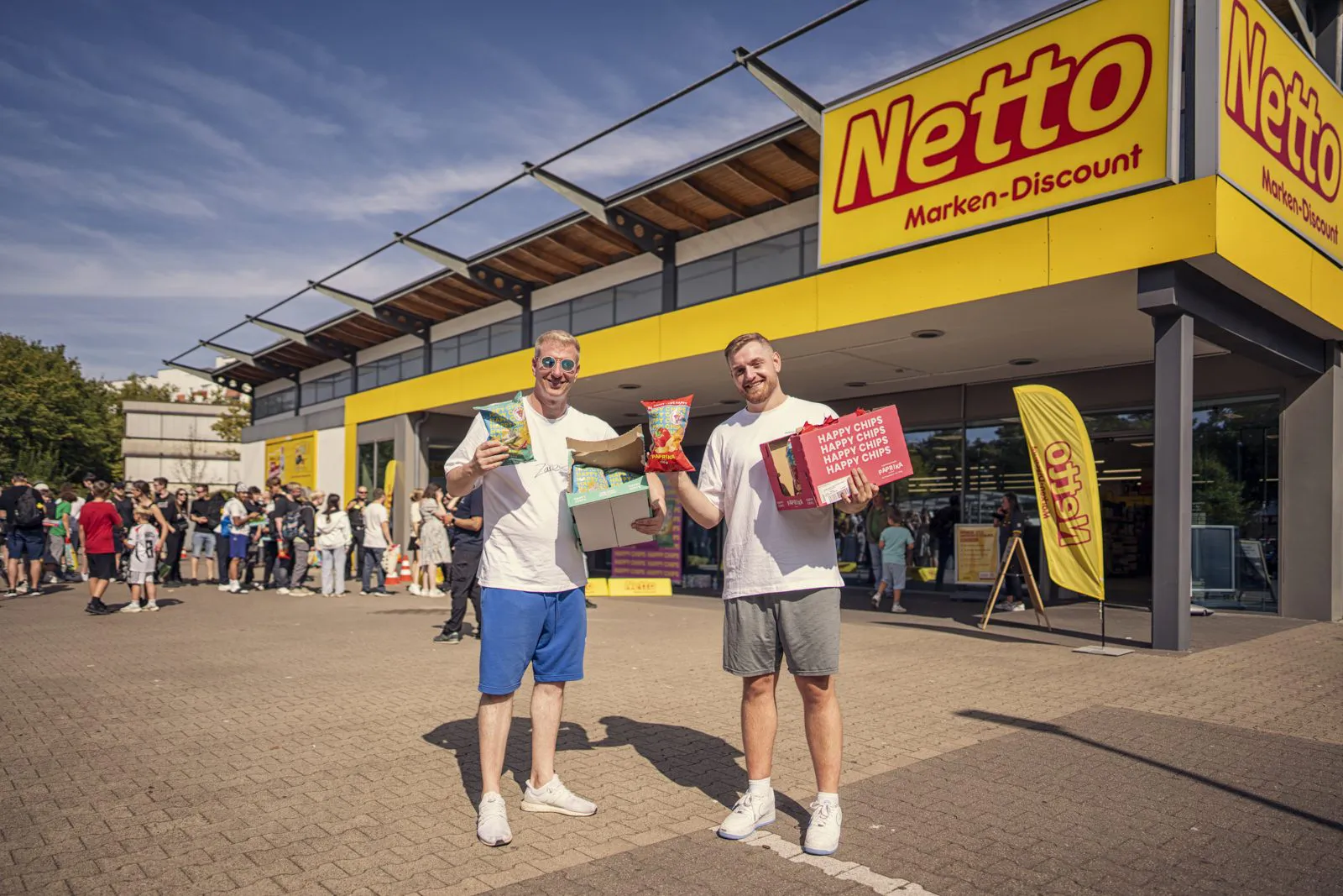 Zwei Männer präsentieren Happy-Chips-Tüten und Kartons vor einer Netto-Filiale, dahinter Wartende im Sonnenlicht.