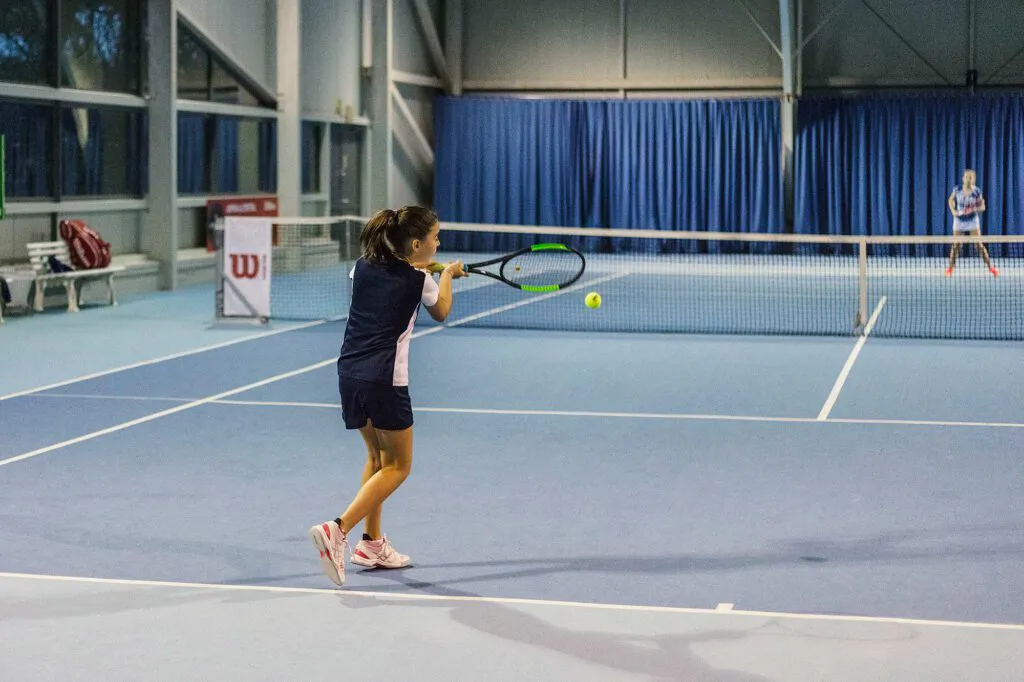 Tennisspielerin schlägt beim Wilson Master Cup einen Ball in einer beleuchteten Halle mit blauem Boden.